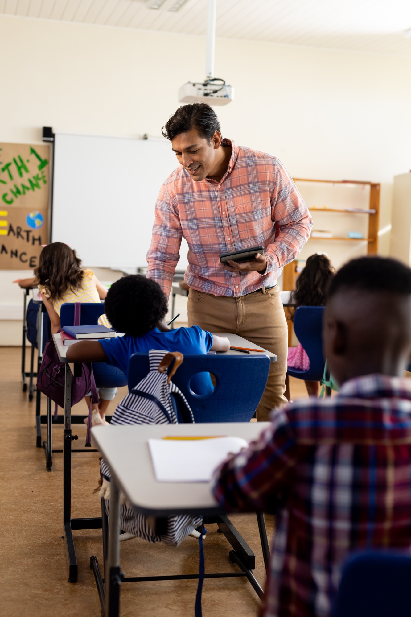Diverse male teacher with tablet and elementary schoolchildren sitting at desks in class
